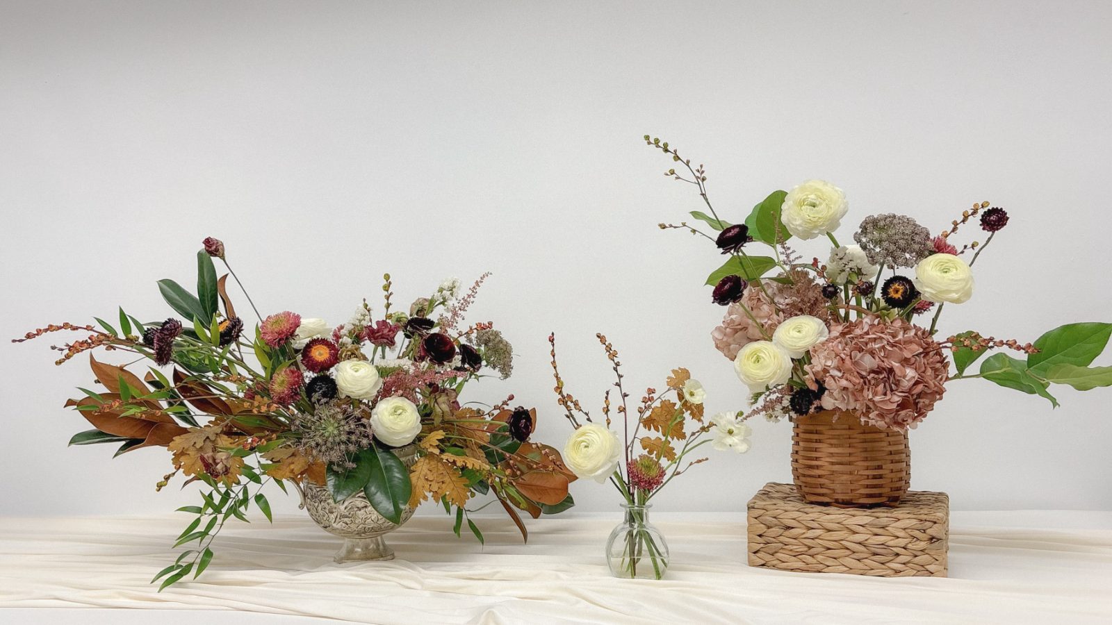 A low arrangement in a silver footed bowl, a single bud vase, and a tall arrangement in a woven basket on a cream cloth
